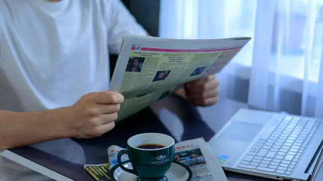 Osoba czyta papierową gazetę przy biurku, obok stoi otwarty laptop i kubek kawy