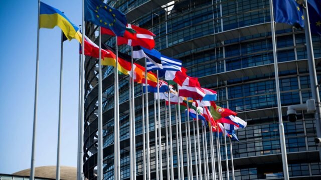 Flagi państw Unii Europejskiej przed fasadą nowoczesnego budynku.