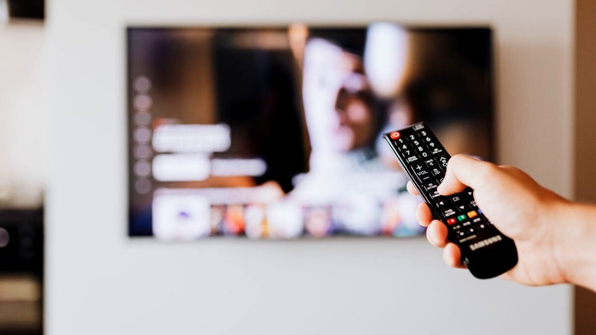 Ręka trzyma pilota przed telewizorem w jasnym pokoju, ekran nieostry