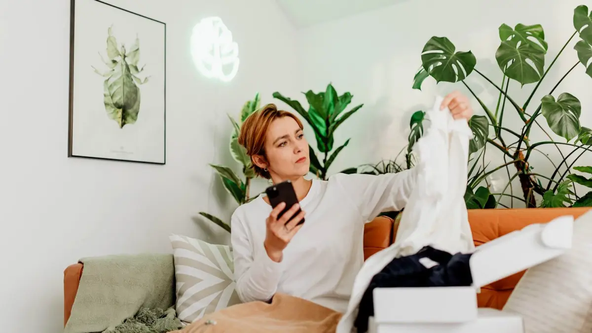 Kobieta w domu robi zakupy na smartfonie, trzymając telefon w obu dłoniach.