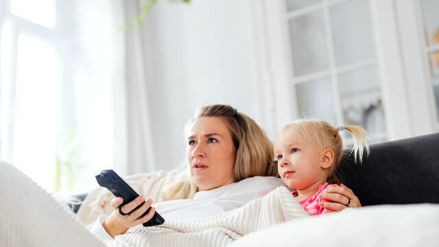 Mama z małą córką na kanapie ogląda telewizję, w ręku trzyma pilota