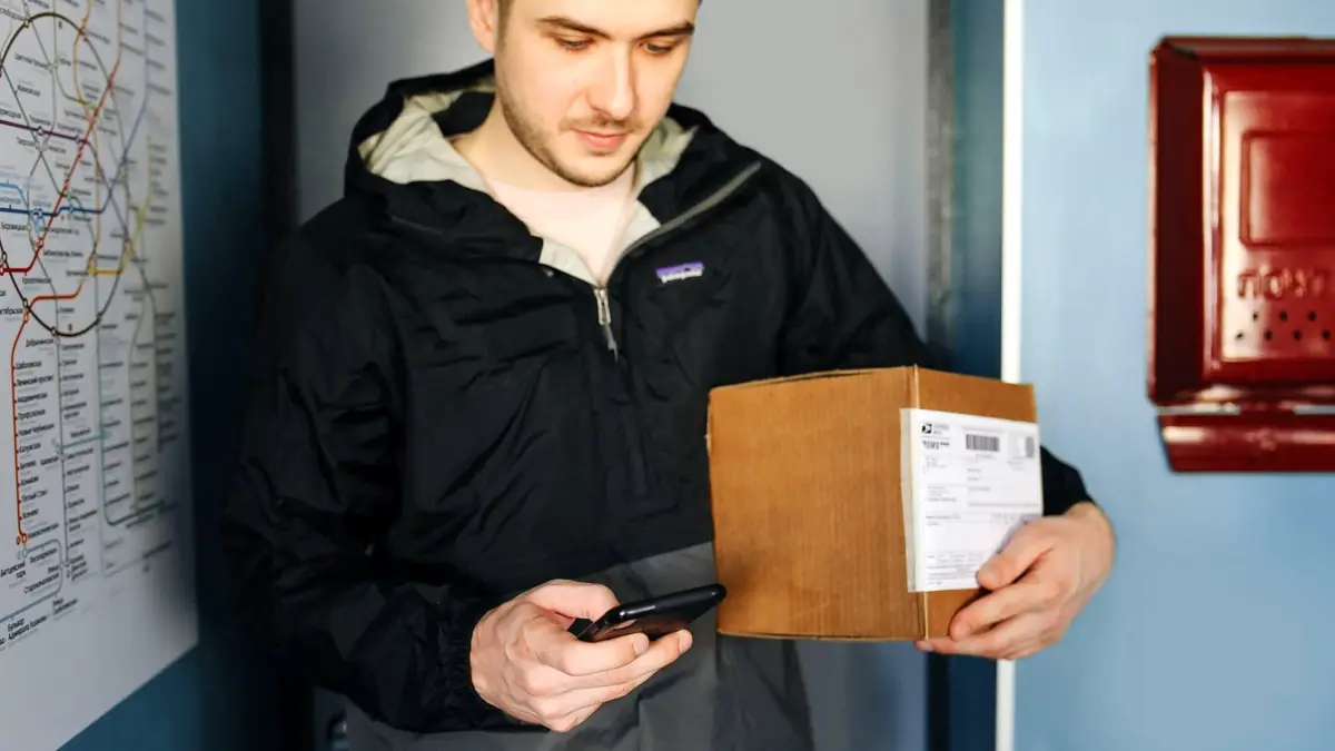 Osoba trzyma smartfon z otwartą aplikacją przed automatem paczkowym na osiedlowej ulicy.