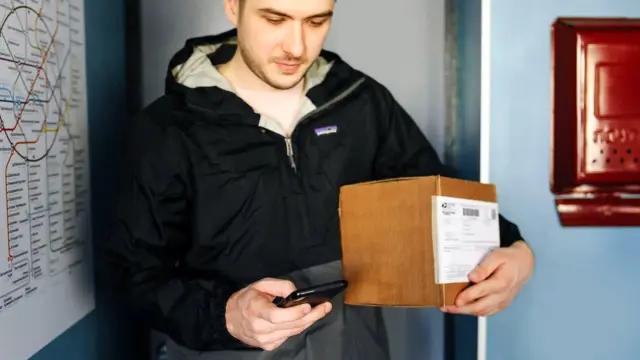 Osoba trzyma smartfon z otwartą aplikacją przed automatem paczkowym na osiedlowej ulicy.