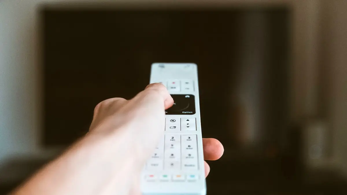 Zbliżenie dłoni z pilotem skierowanym w stronę telewizora we wnętrzu.