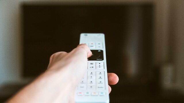 Zbliżenie dłoni z pilotem skierowanym w stronę telewizora we wnętrzu.
