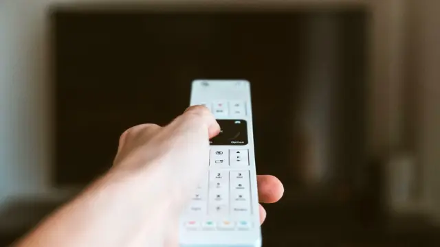 Zbliżenie dłoni z pilotem skierowanym w stronę telewizora we wnętrzu.