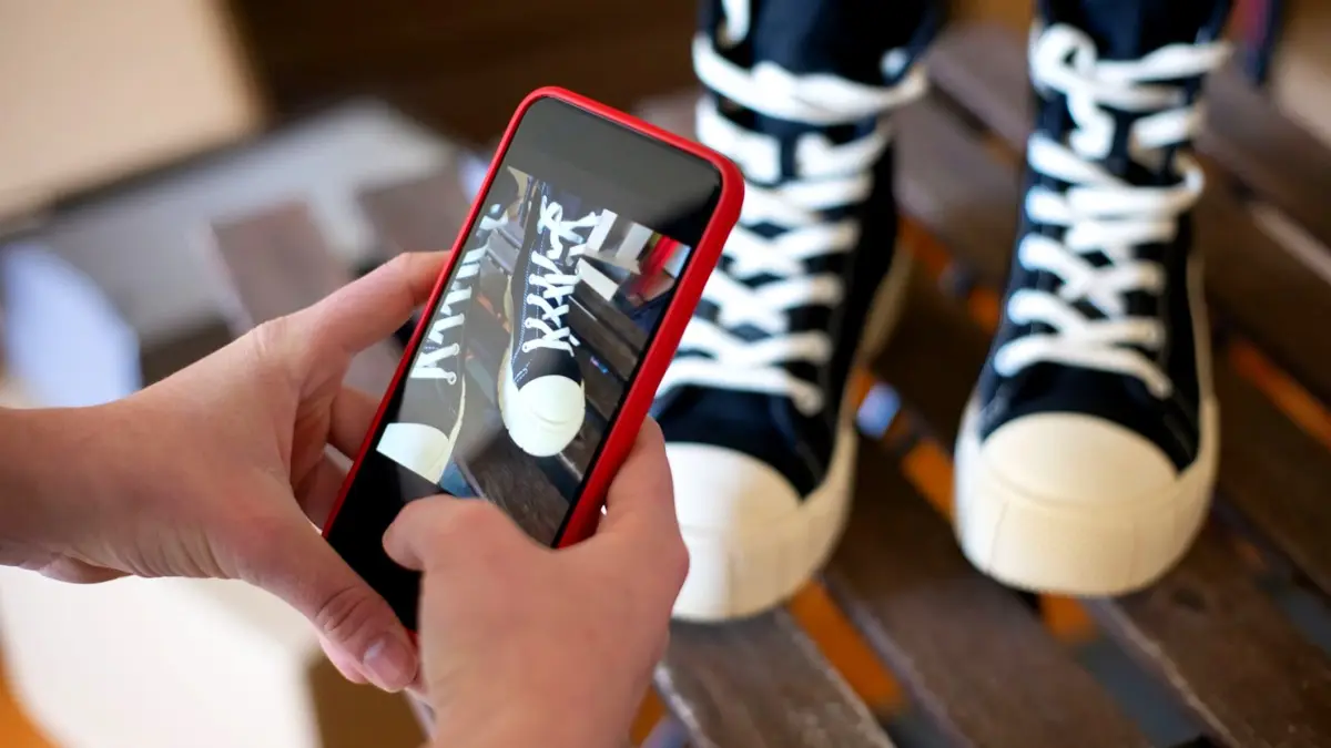 Dłonie trzymające smartfon, który fotografuje czarne sneakersy leżące na jasnym blacie.
