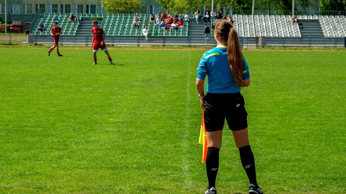 Sędzia piłkarska prowadzi mecz na stadionie, w tle murawa i zawodnicy