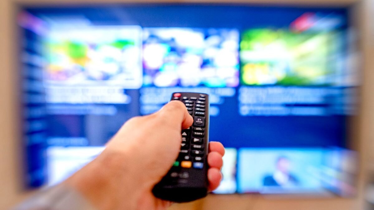 Dłoń z pilotem skierowanym w stronę rozmytego ekranu telewizora.
