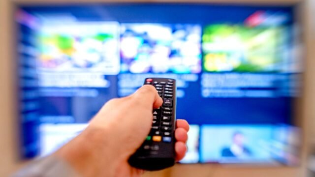 Dłoń z pilotem skierowanym w stronę rozmytego ekranu telewizora.
