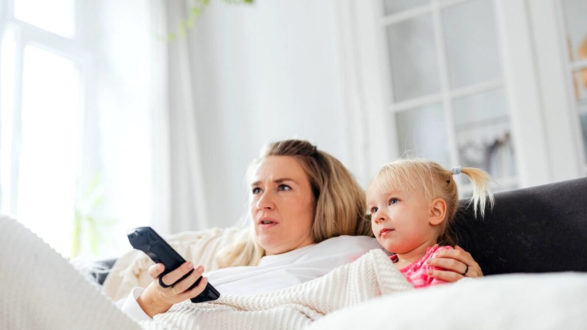 Mama i córka na kanapie w jasnym salonie, jedna trzyma pilot do telewizora