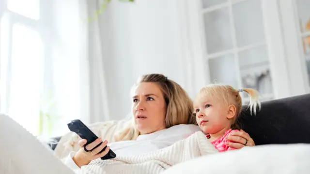 Mama i córka na kanapie w jasnym salonie, jedna trzyma pilot do telewizora