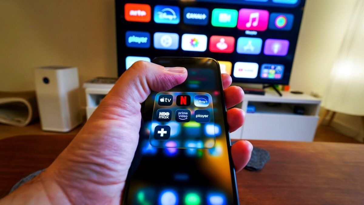 Zbliżenie dłoni z telefonem, na ekranie aplikacje streamingowe, w tle telewizor z ikonami serwisów.