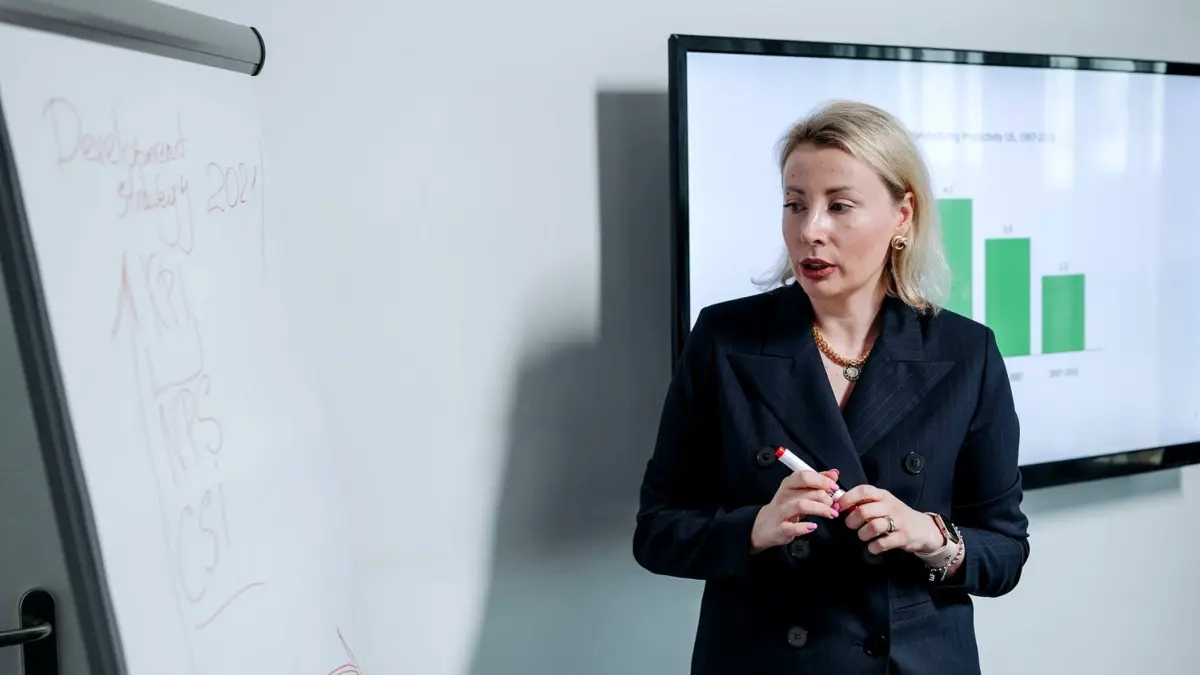 Kobieta przy tablicy omawia wykres słupkowy wyświetlony na ekranie w sali konferencyjnej