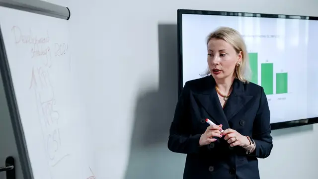 Kobieta przy tablicy omawia wykres słupkowy wyświetlony na ekranie w sali konferencyjnej