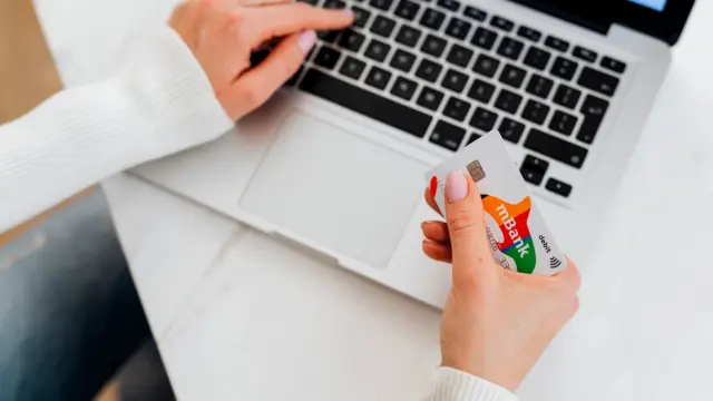 Zbliżenie na dłonie przy laptopie, jedna trzyma kartę płatniczą nad klawiaturą