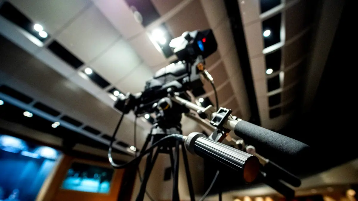 Kamera telewizyjna na statywie w studiu, zbliżenie na obiektyw i ekran podglądu.