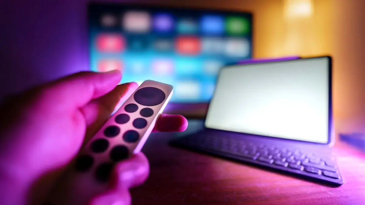 Dłoń z pilotem przed telewizorem i laptopem w pokoju