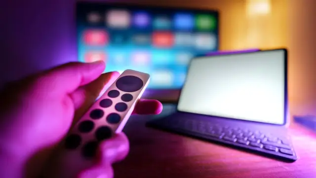 Dłoń z pilotem przed telewizorem i laptopem w pokoju