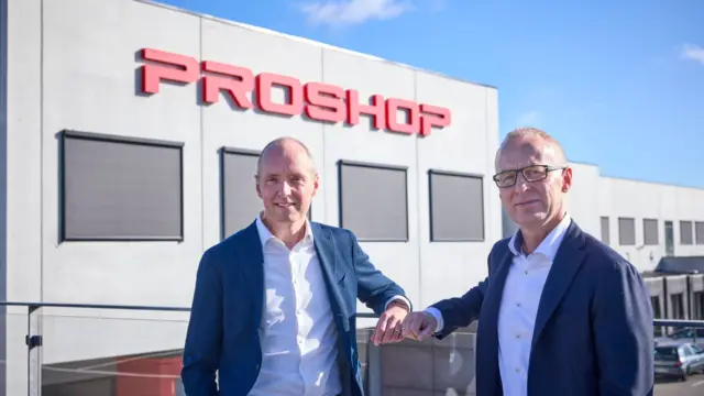 Dwóch mężczyzn w garniturach przed budynkiem z dużym czerwonym napisem Proshop