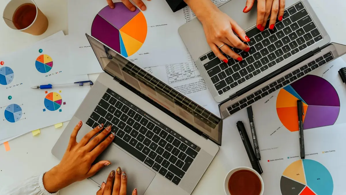 Dwie osoby przy laptopach nad wydrukami wykresów kołowych i słupkowych, widok z góry.