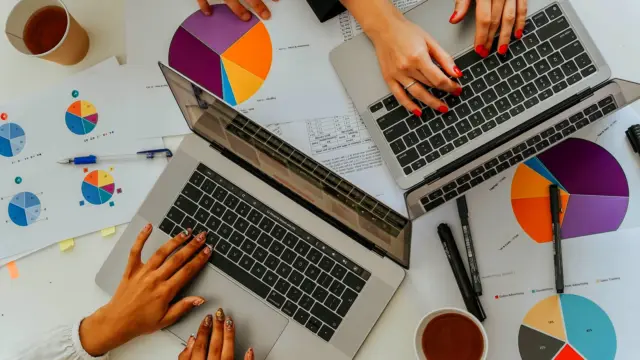 Dwie osoby przy laptopach nad wydrukami wykresów kołowych i słupkowych, widok z góry.